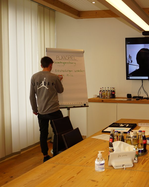 Junge schreibt auf Flipchart, anderer Junge sitzt am Besprechungstisch, zwei Personen per Teams Meeting zugeschaltet | © RÖSLE GROUP