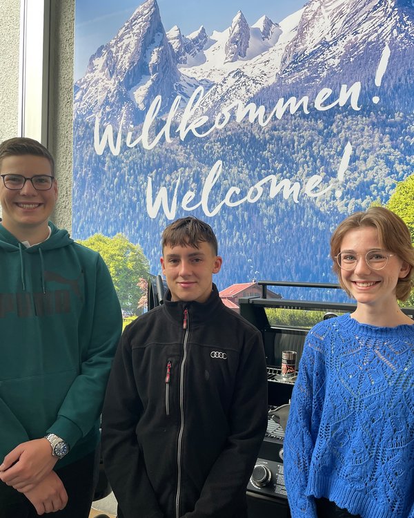 Ausbildungsbeginn 2023 | © RÖSLE GROUP zwei Jungen und ein Mädchen stehen lächelnd vor einem "Welcome"-Plakat mit Bergmotiv | © RÖSLE GROUP
