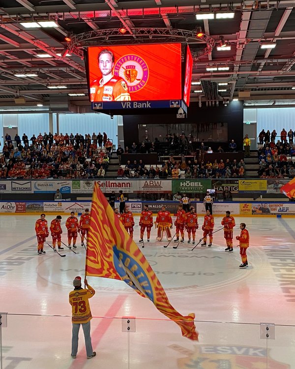 Eishockey-Spieler stehen im Halbkreis auf der Eisfläche im vollen Stadion, ein Mann auf der Eisfläche hält eine Fahne des Vereins in der Hand | © RÖSLE GROUP