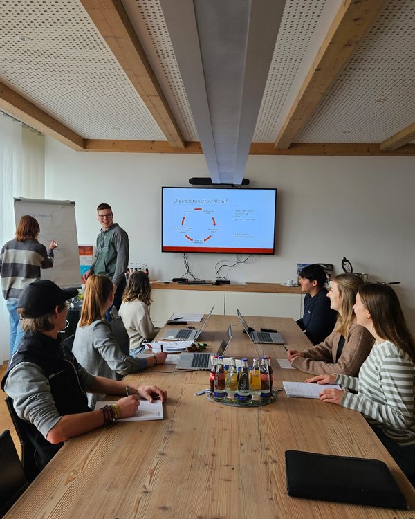 Planspiel 2024 | © RÖSLE GROUP Sechs Auszubildende sitzen mit Unterlagen und Laptops an einem Tisch, zwei weitere schreiben etwas auf ein Flipchart | © RÖSLE GROUP