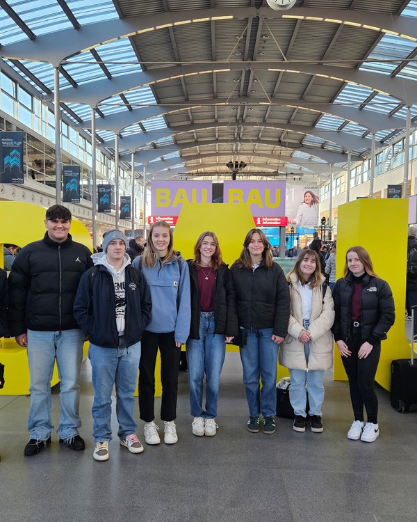 Besuch Messe BAU 2025 | © RÖSLE GROUP Fünf Mädchen und drei Jungen, die vor großen, gelben BAU-Buchstaben auf der Messe BAU in München stehen | © RÖSLE GROUP